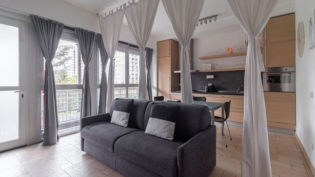 A modern living room with a grey sofa, a dining table, and a kitchen area with wooden cabinets and stainless steel appliances. - Italianway Comfort - Savona 123 (Milan)