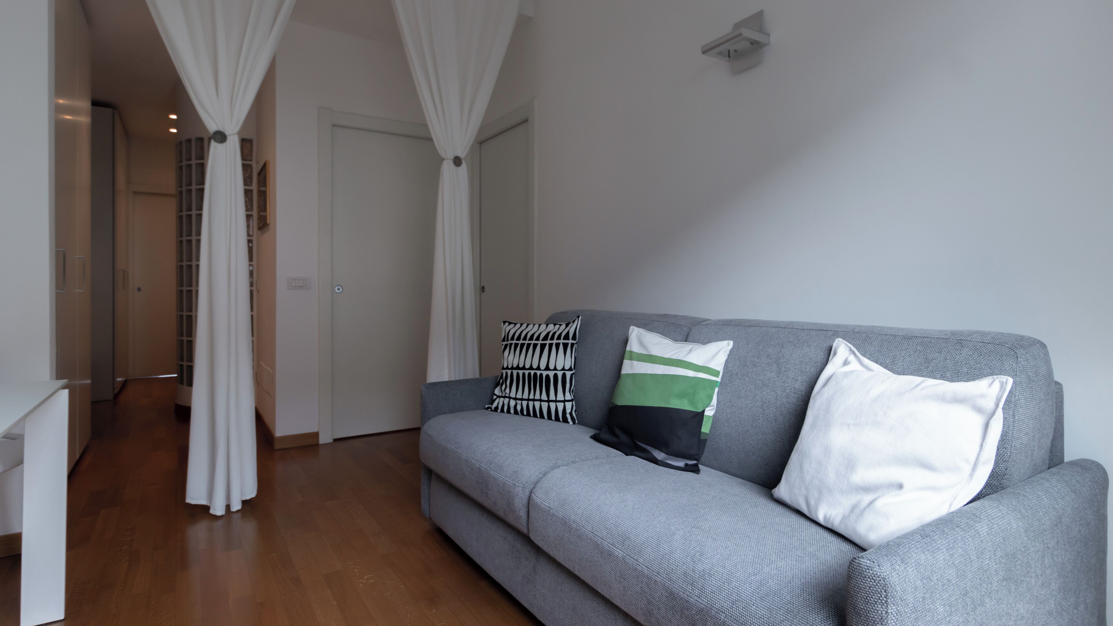 Een moderne woonkamer met een grijze bank, witte gordijnen en een houten vloer.