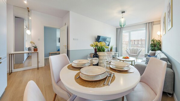 A dining area with a round table set for four, a kitchen in the background, and a city view through large windows. - Holiday City (Kolobrzeg)