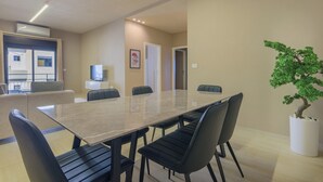 A modern dining area with a long table, black chairs, a potted tree, and a view of the outside.