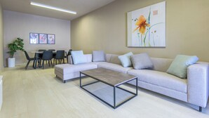 A modern living room with a sectional sofa, a coffee table, and a dining area with chairs and a table. There is a large painting on the wall and a potted plant.