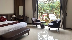Habitación de hotel con cama, escritorio, dos sillones, un banco, lámpara y vista a zonas verdes a través de un ventanal decorativo.
