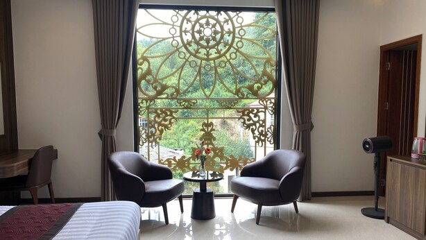 Habitación de hotel con cama, dos sillones, una mesita con flores, una vista decorativa desde la ventana y un escritorio.