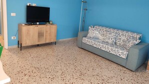 A living room with a blue wall, a grey sofa, a wooden cabinet with a TV, and a floor lamp. - Turiddu Apartment- Casa Vacanze Giardini Naxos (Taormina)