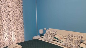 A neatly made bed with a green bedspread and multiple pillows, a bedside table with a lamp, and a patterned curtain. - Turiddu Apartment- Casa Vacanze Giardini Naxos (Taormina)