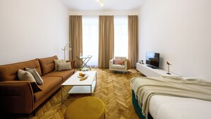 Una habitación de hotel moderna con sofá, una mesita, una silla, cama, televisión y ventanas grandes con cortinas.