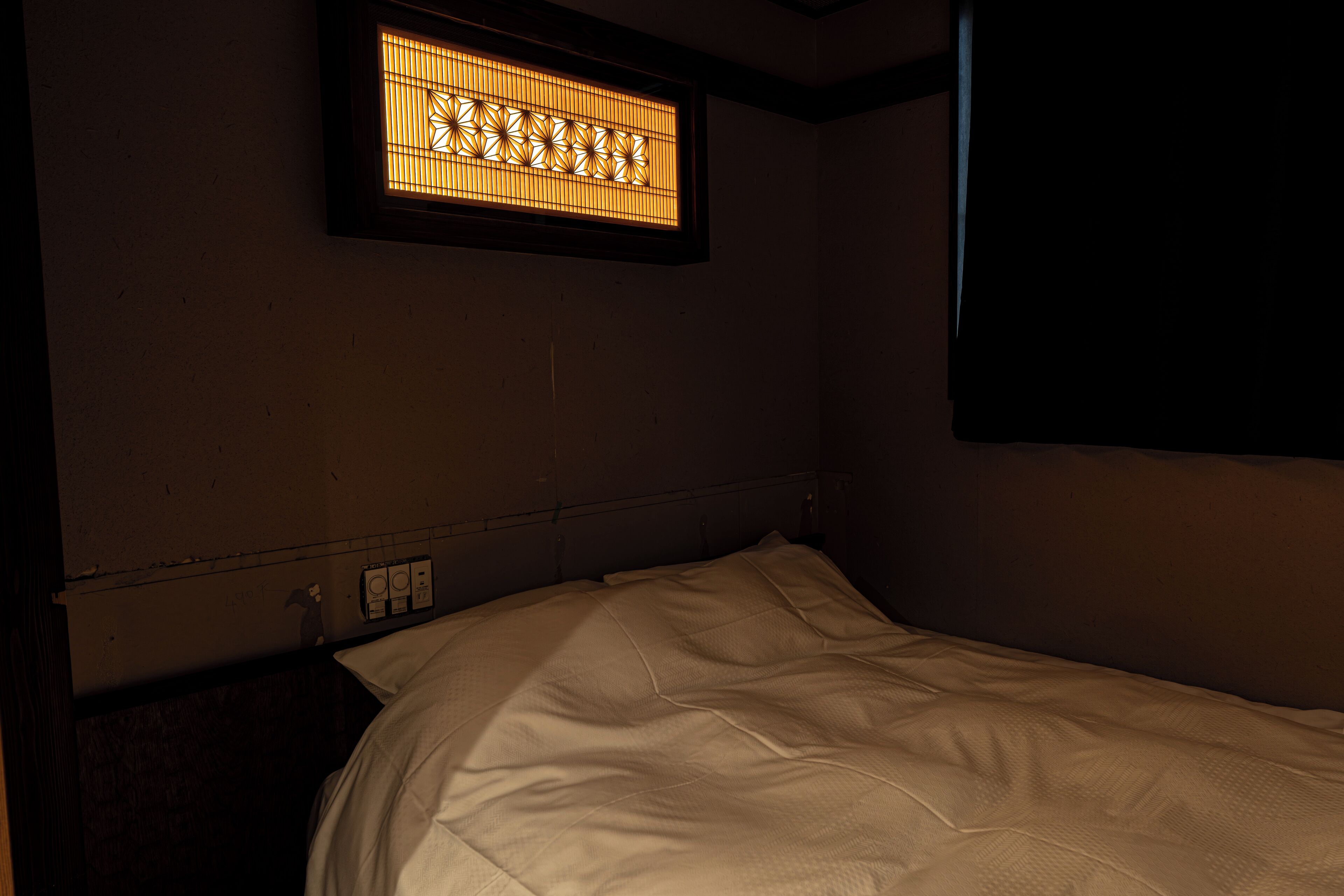A single bed with a headboard, a wall-mounted light fixture, and a power outlet.