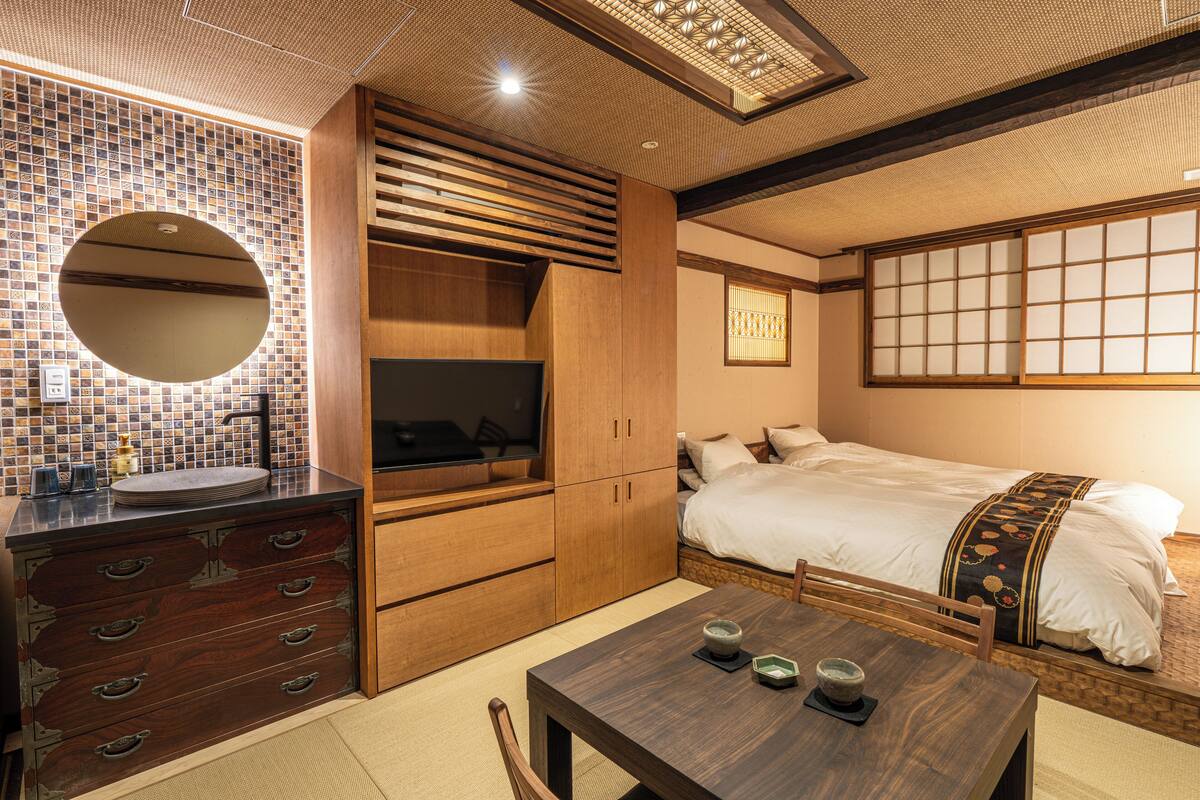 A traditional Japanese-style room with a tatami mat floor, a wooden dresser with drawers, a round mirror, a bed with a white bedspread and a decorative headboard, and a small table with a tea set.