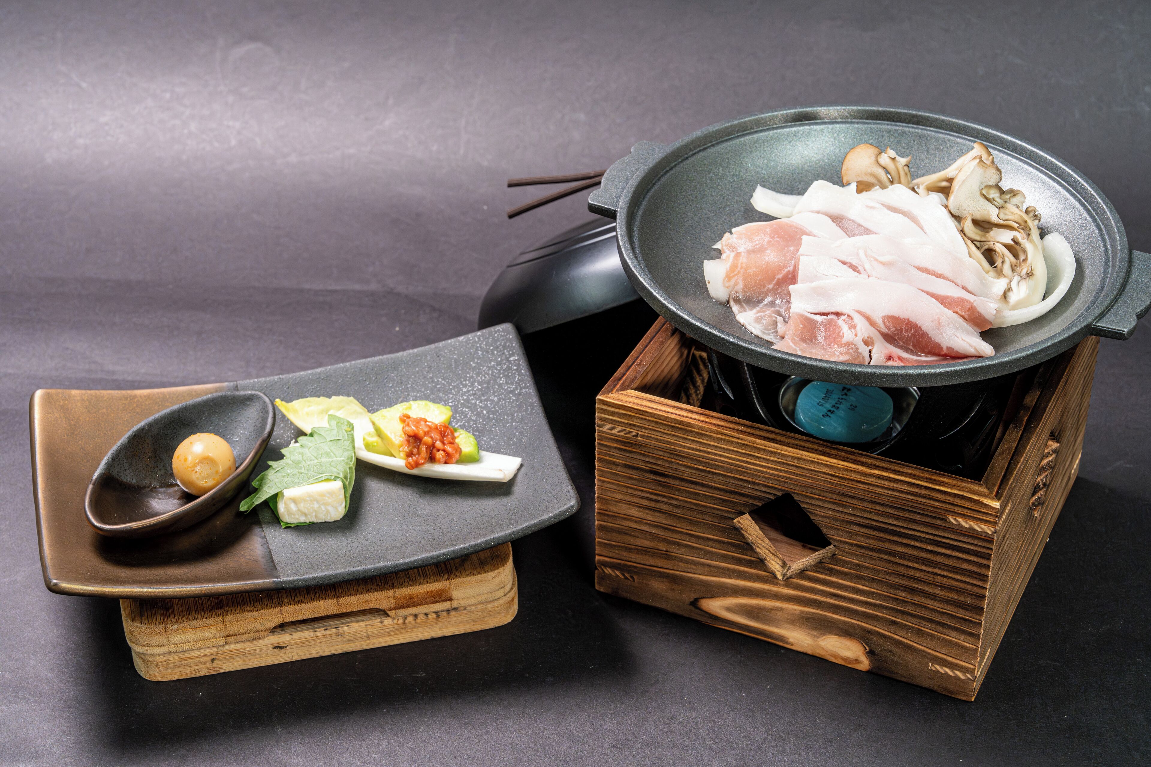 A wooden box with a metal pan containing sliced meat and mushrooms, a small dish with a ball of food and greens, and a black object with chopsticks.