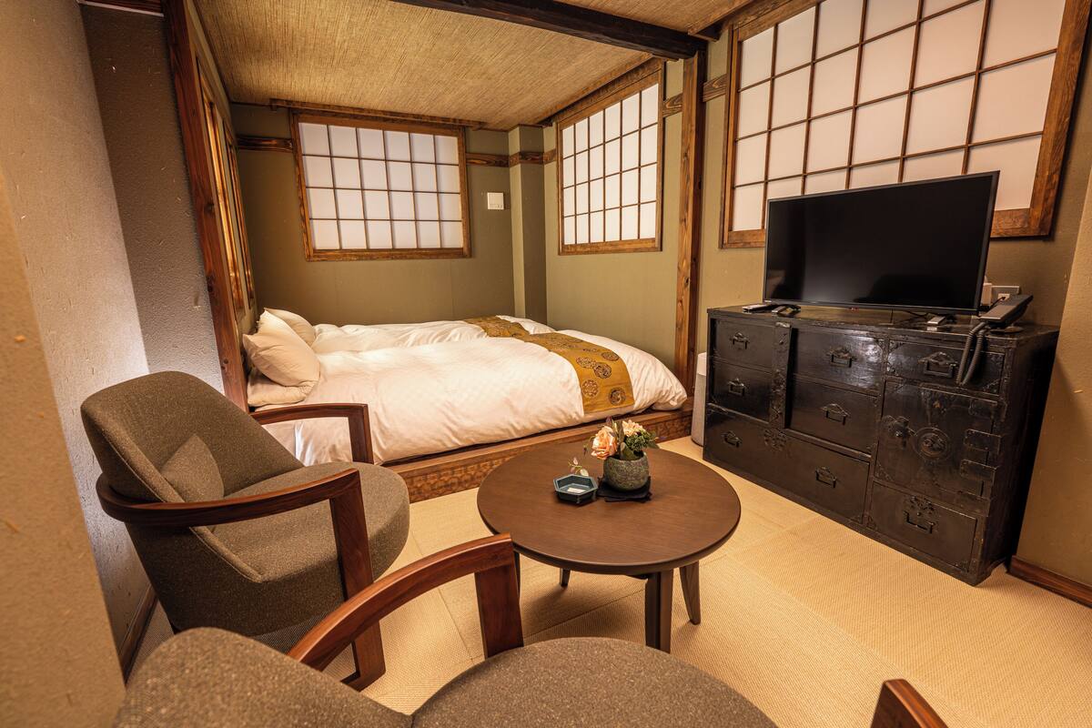 A traditional Japanese room with tatami flooring, a bed with a white and gold bedspread, a television on a wooden cabinet, and a small table with a vase.