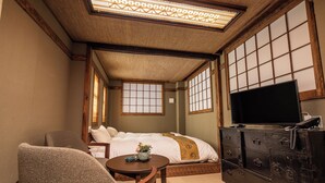 Une chambre de style japonais traditionnel, avec un sol recouvert de tatamis, un futon servant de lit, un téléviseur posé sur une commode et une petite table sur laquelle se trouve un vase.