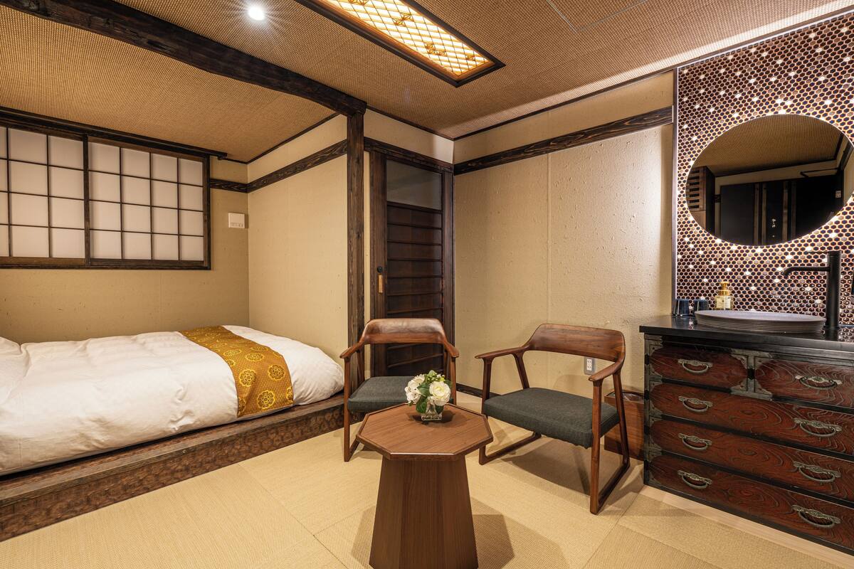 A traditional Japanese room with tatami flooring, a low wooden bed, two chairs, a small round table with a vase, and a dresser with drawers.