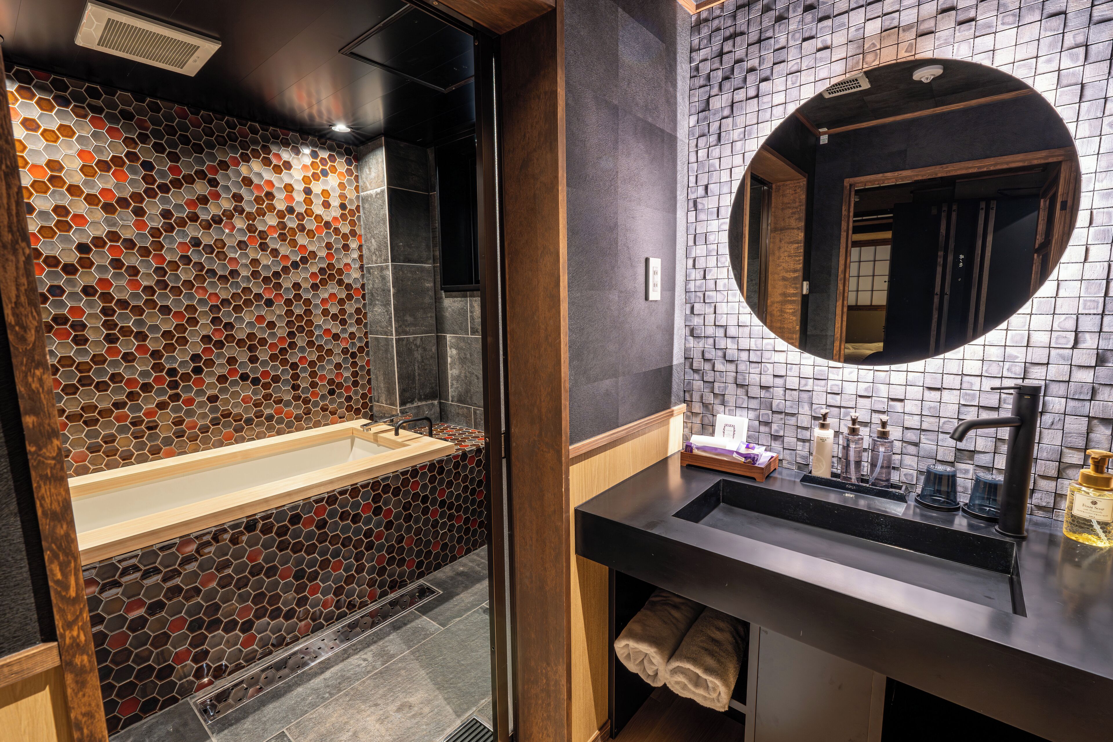 Une salle de bain moderne avec un revêtement mural en carreaux hexagonaux, une baignoire sur pieds, un miroir rond et un plan de toilette noir doté d’un lavabo et d’accessoires de toilette.