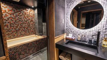 A modern bathroom with a hexagonal tile backsplash, a freestanding bathtub, a round mirror, and a black countertop with a sink and toiletries.