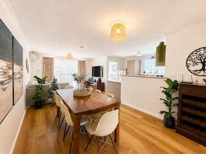 Una sala de estar moderna con una mesa de comedor de madera, sillas blancas, un televisor y una zona de cocina.
