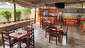 A restaurant with wooden tables and chairs, a thatched roof, and a kitchen area with a sign reading "Seagull Mangwiresort". - Seagull Nungwi Resort (Nungwi)