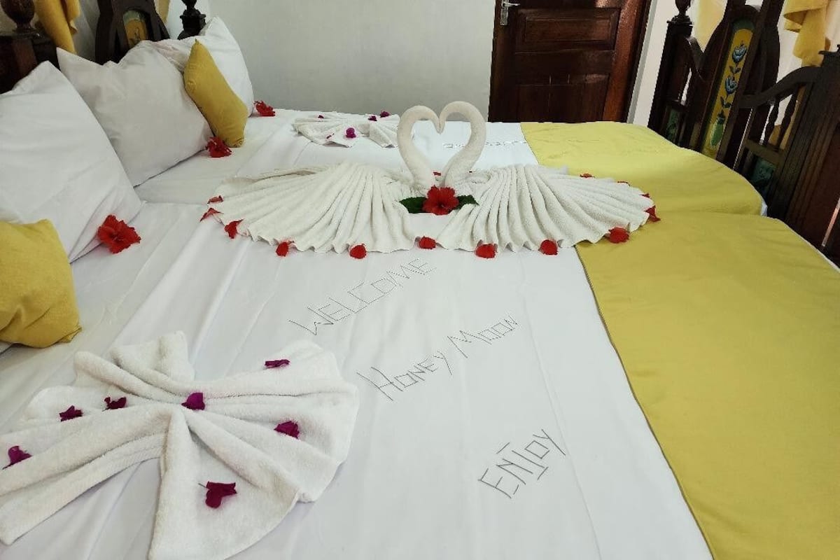 A bed with a white sheet, yellow pillows, and a heart-shaped towel arrangement with red flowers. Text on the bed reads "HELLO, ENJOY."