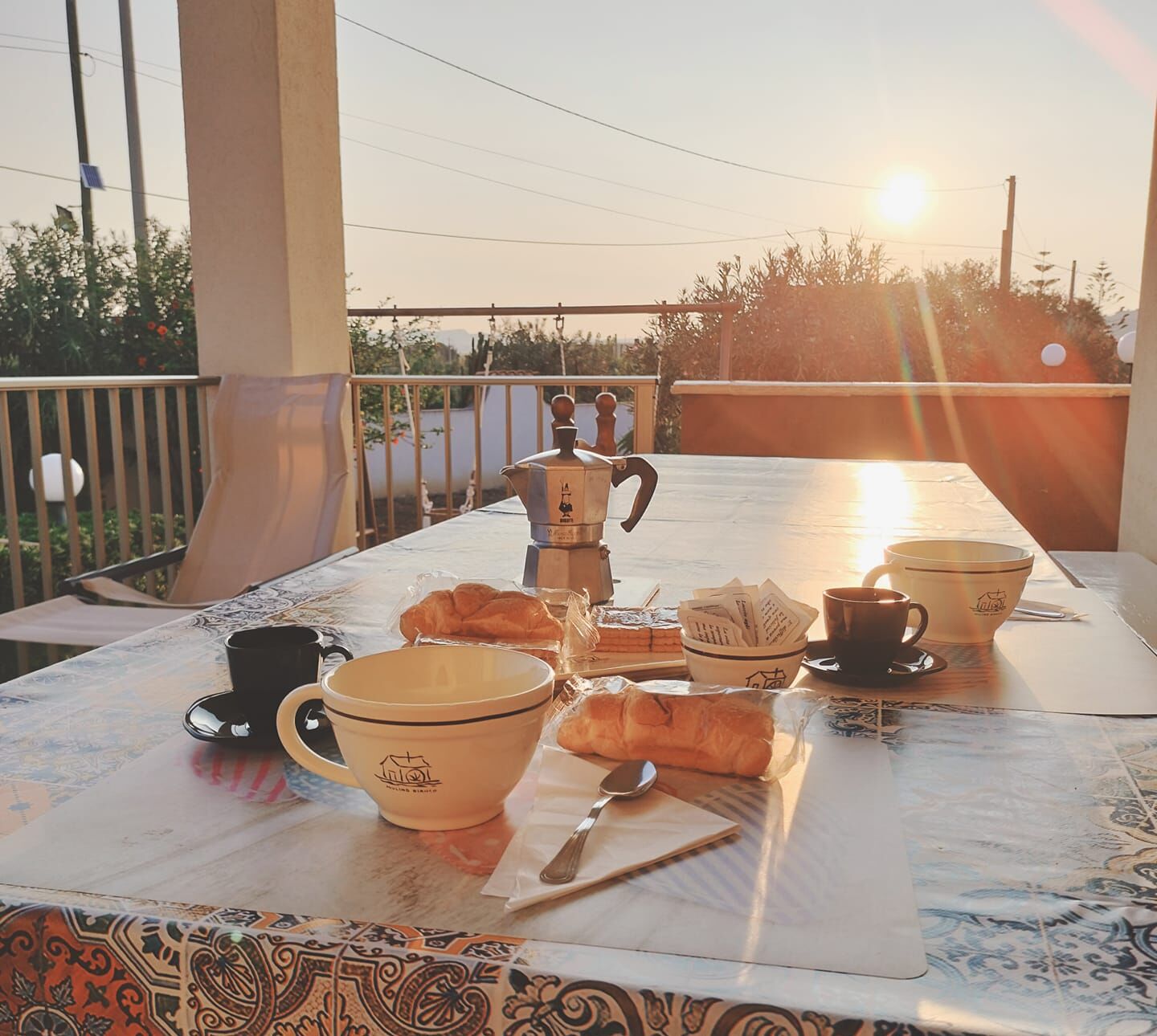 A breakfast setup on a balcony with a table, coffee maker, cups, and pastries.