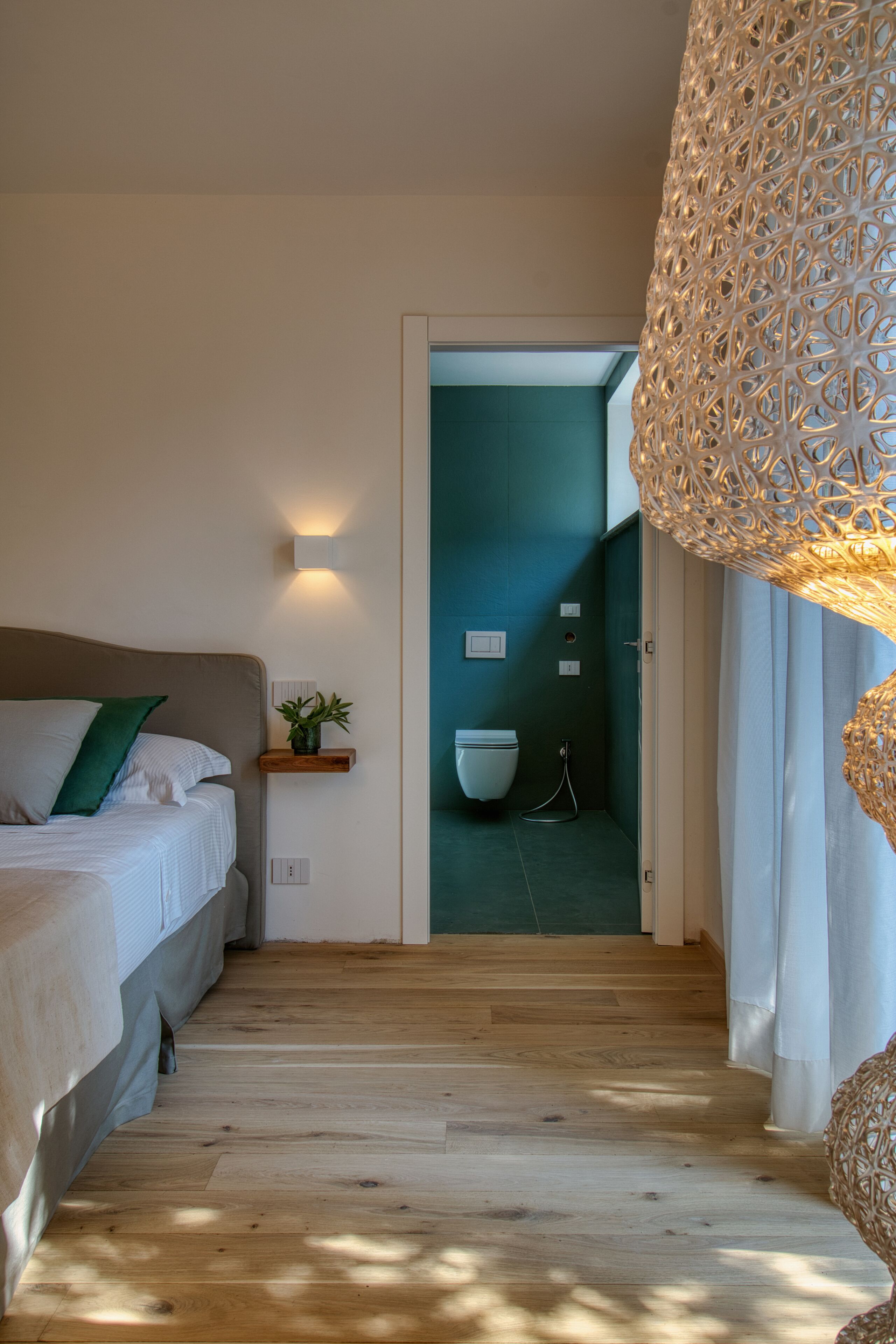 A bedroom with a wooden floor, a bed with white bedding, a green pillow, a bedside plant, and a wall-mounted light fixture.