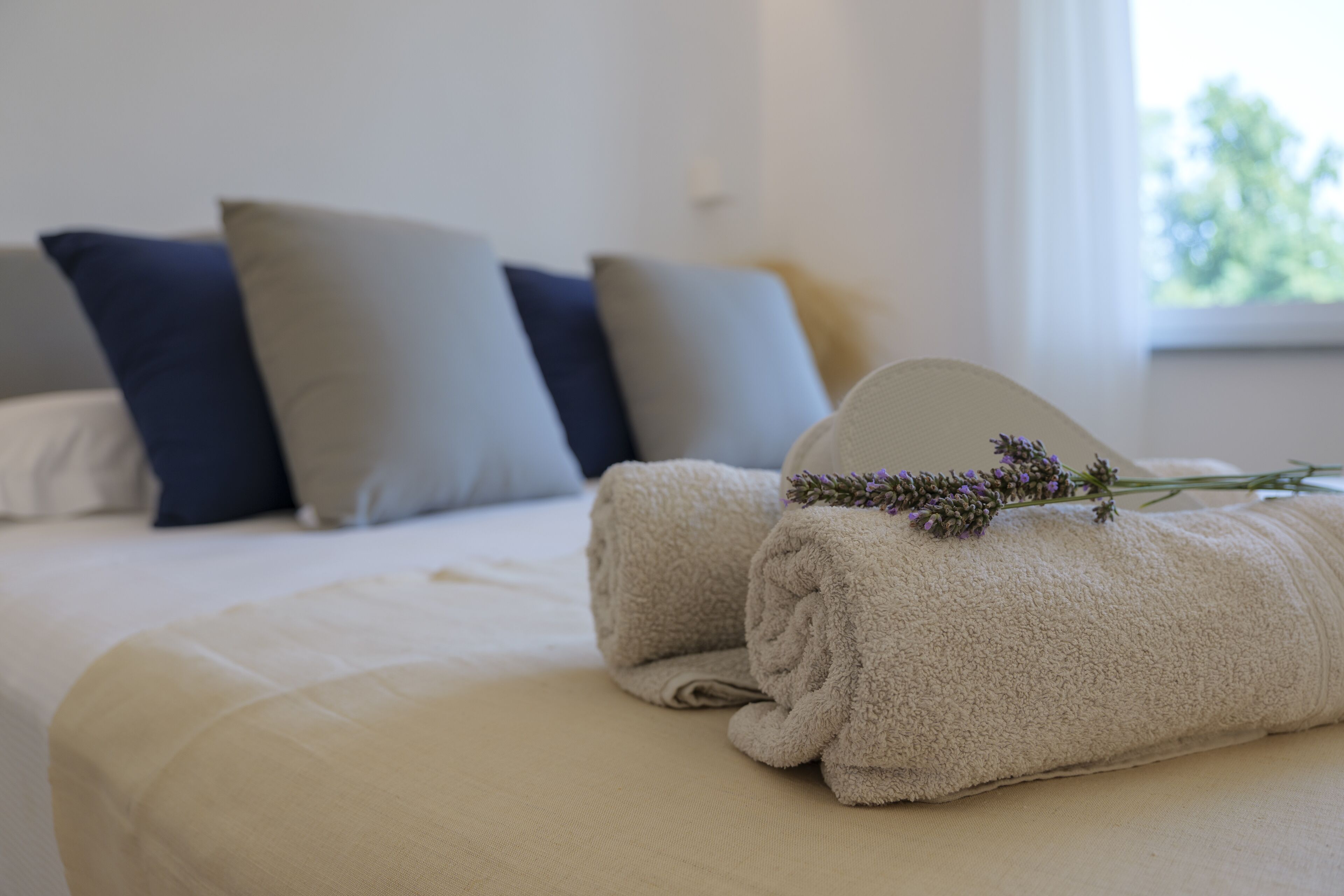 A neatly made bed with two rolled towels and a lavender sprig on top.