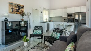 Un salon moderne avec un canapé gris, des fauteuils en cuir noir et une table basse en verre. On y trouve également un coin repas avec une table et des chaises blanches, ainsi qu’une cuisine dotée de meubles blancs et d’appareils électroménagers en acier inoxydable.