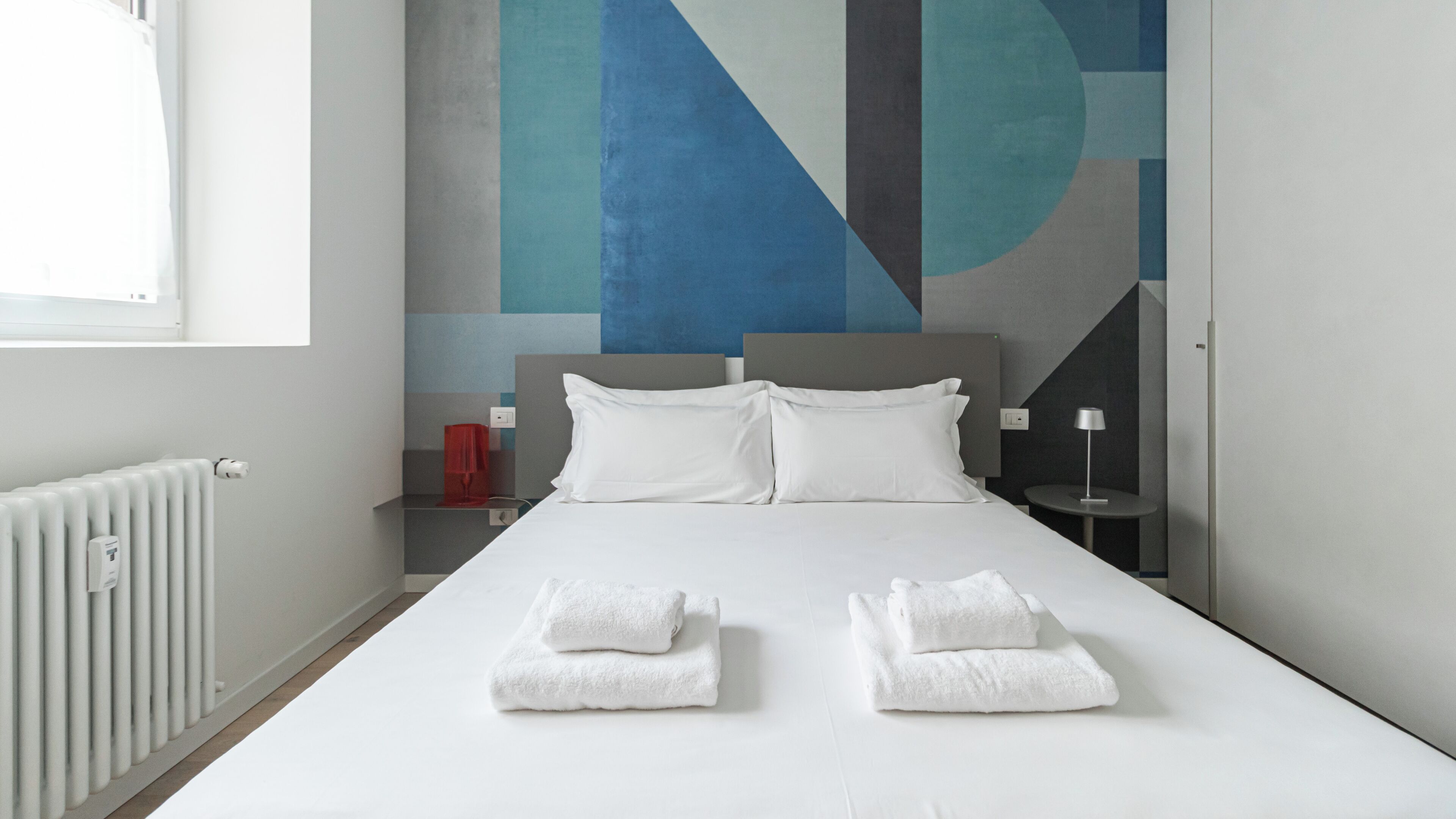 A modern hotel room with a large bed, white linens, and a geometric wall mural.