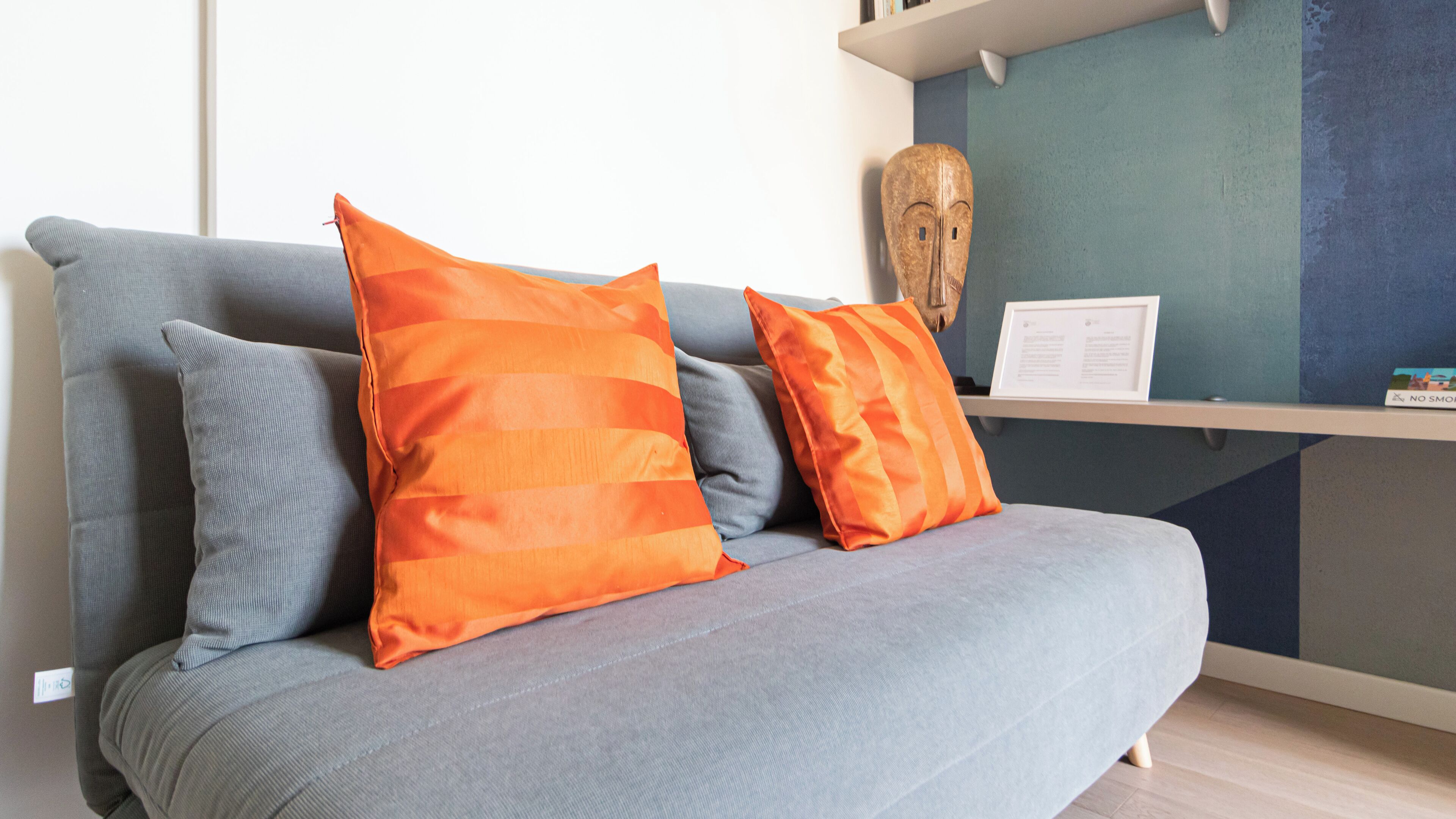 A grey sofa with two orange pillows and a wooden mask on a shelf.