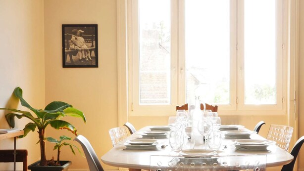 Un coin repas avec une table ronde dressée pour six personnes, une plante en pot et un tableau encadré accroché au mur.