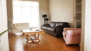 Un salon avec un plancher en bois, un canapé en cuir marron, un fauteuil blanc, un fauteuil rose, une table basse et une lampe.