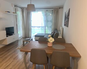 A modern living room with a sofa, a dining table with chairs, a TV on a stand, and a large window with curtains. - Cozy apartment with 1 bedroom and AC, WiFi in welcoming Pécs (Pécs)