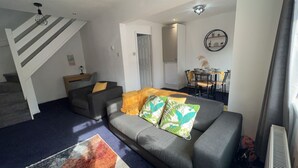 A living room with a sofa, a dining table with chairs, a staircase, and a wall-mounted shelf. - Cozy 2 Bedhouse 20mins From Manchester City Centre (Oldham)