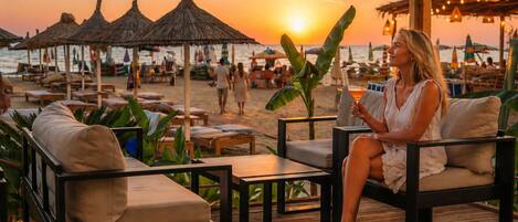 A beachside lounge with outdoor seating, a sunset view, and thatched umbrellas.