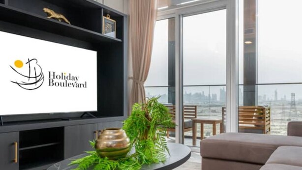 Een moderne woonkamer met een grote flatscreen televisie, een ronde salontafel met een decoratieve plant en een bank. De kamer biedt uitzicht op de stad door grote ramen.