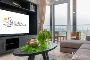 A modern living room with a large flat-screen TV, a round coffee table with a decorative plant, and a sofa. The room has a city view through large windows.