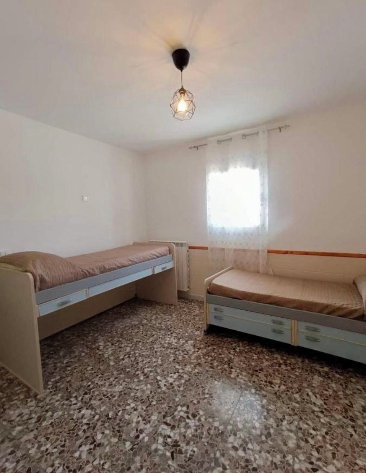 A small, single-room apartment with two beds, a ceiling light, a window with curtains, and a speckled floor.