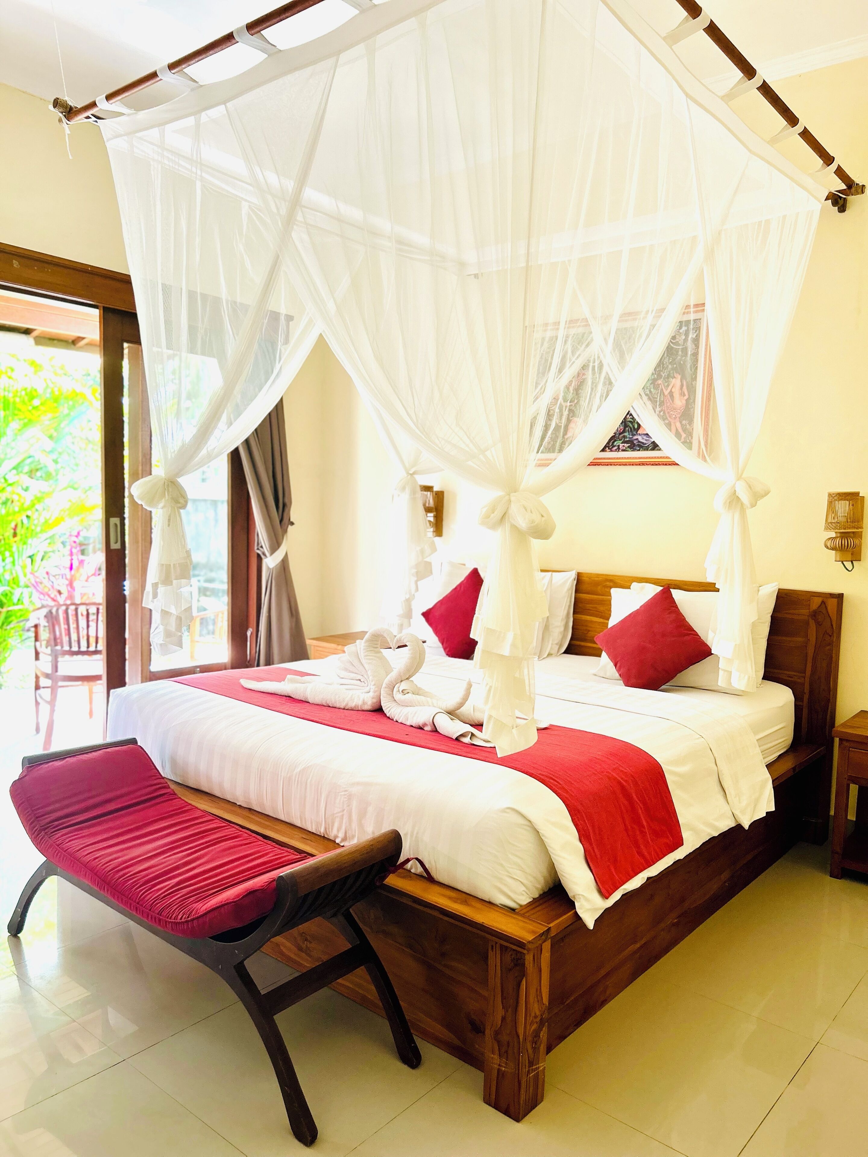 Una cama con dosel y cuatro columnas con un edredón rojo y blanco, un banco de madera y vistas al exterior con vegetación.