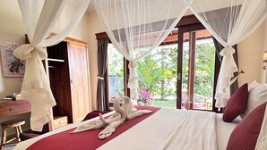A bedroom with a bed, a desk, and a window with a view of greenery. - Canting Bali Suite (Ubud)