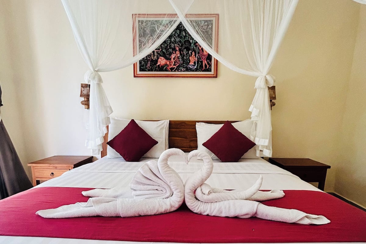 A hotel room with a bed, a nightstand, a wooden headboard, a painting on the wall, and a canopy over the bed.