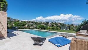En swimmingpool med liggestole, udsigt over en bakke med huse, og en klar blå himmel.