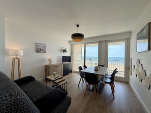 A modern living room with a dining area, large windows with a view of the sea, a flat-screen TV, and a floor lamp.