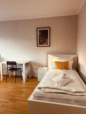 A bedroom with a bed, a desk, a chair, a nightstand, and a framed picture on the wall.