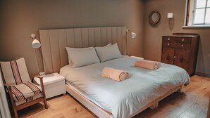Quarto com uma cama grande, duas luminárias de cabeceira, uma mesinha de cabeceira de madeira, uma cadeira e um guarda-roupa de madeira.