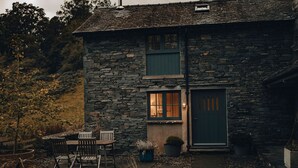 Uma casa de pedra com porta verde, um pequeno alpendre com cadeiras e um vaso de planta.