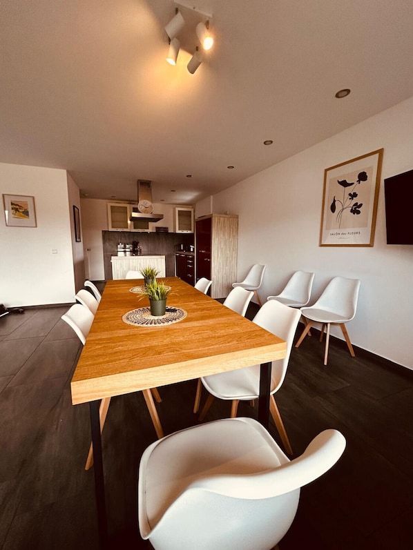 A modern dining area with a wooden table, white chairs, a potted plant, and a kitchen in the background.