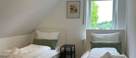 Una habitación pequeña y bien iluminada con dos camas individuales, una mesita y una ventana con vistas a zonas verdes.