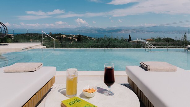 Ein Poolbereich mit Buch, Saftglas und Weinglas auf einem Tisch, mit Blick auf eine malerische Berg- und Wasserlandschaft.