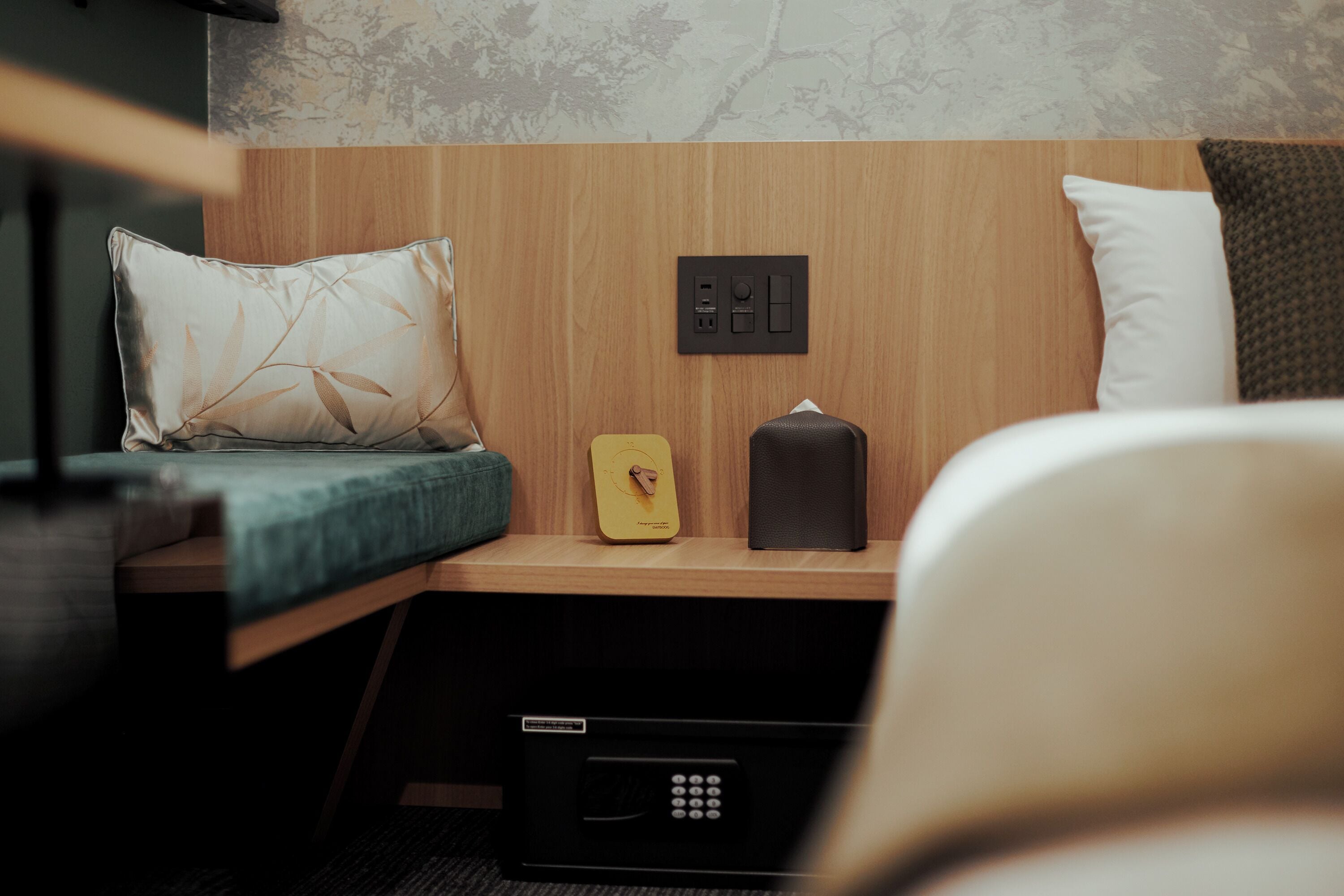 A compact hotel room with a bed, a bedside table with a lamp, a power outlet, and a small storage box.
