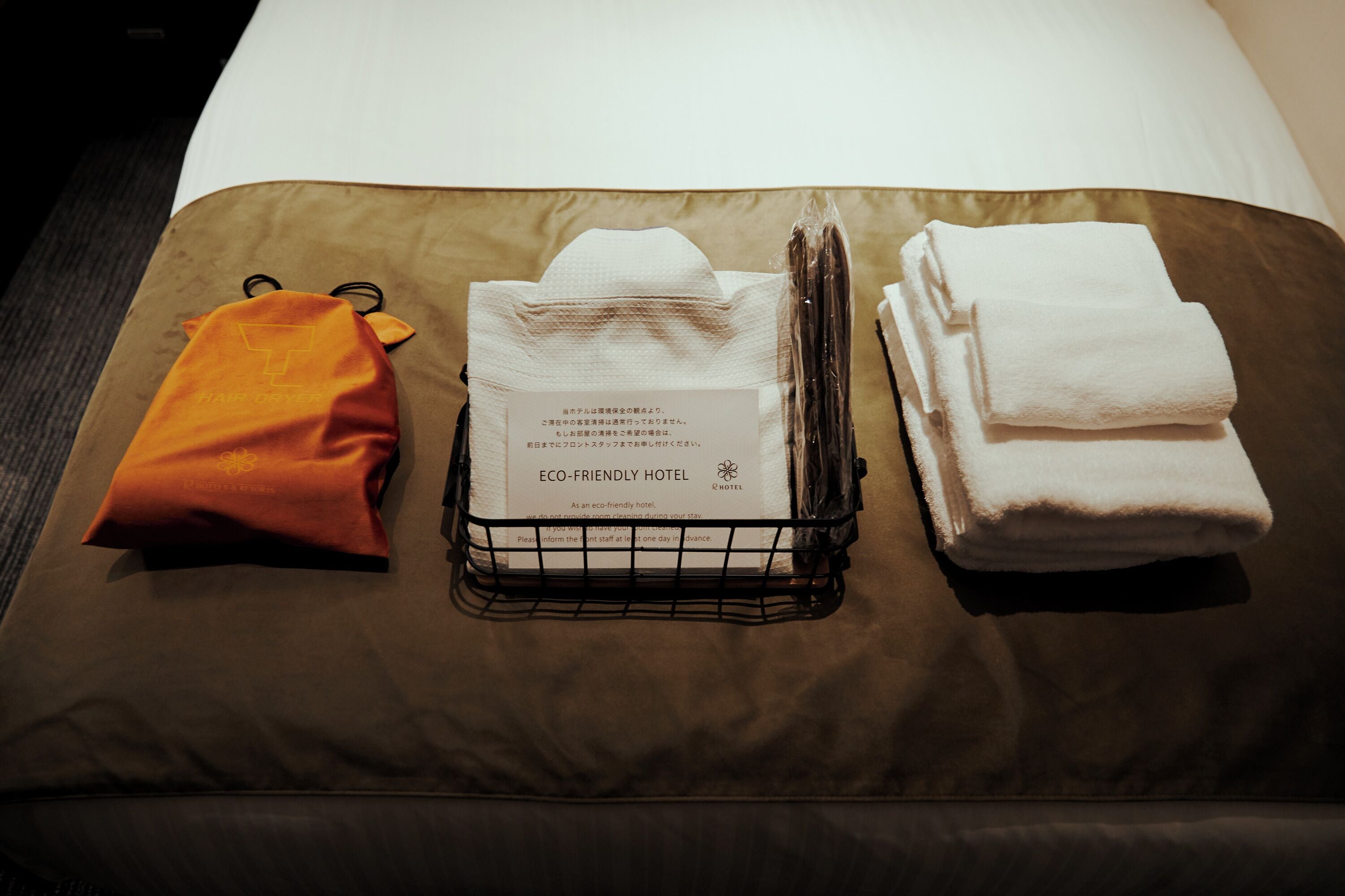 Une chambre d’hôtel bien rangée, avec une couverture marron, un oreiller blanc, une serviette blanche pliée, un petit sac orange et une boîte de mouchoirs portant l’inscription « HÔTEL ÉCOLOGIQUE ».