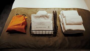 A neatly arranged hotel room with a brown bedspread, a white pillow, a folded white towel, a small orange bag, and a tissue box labeled "ECO-FRIENDLY HOTEL".
