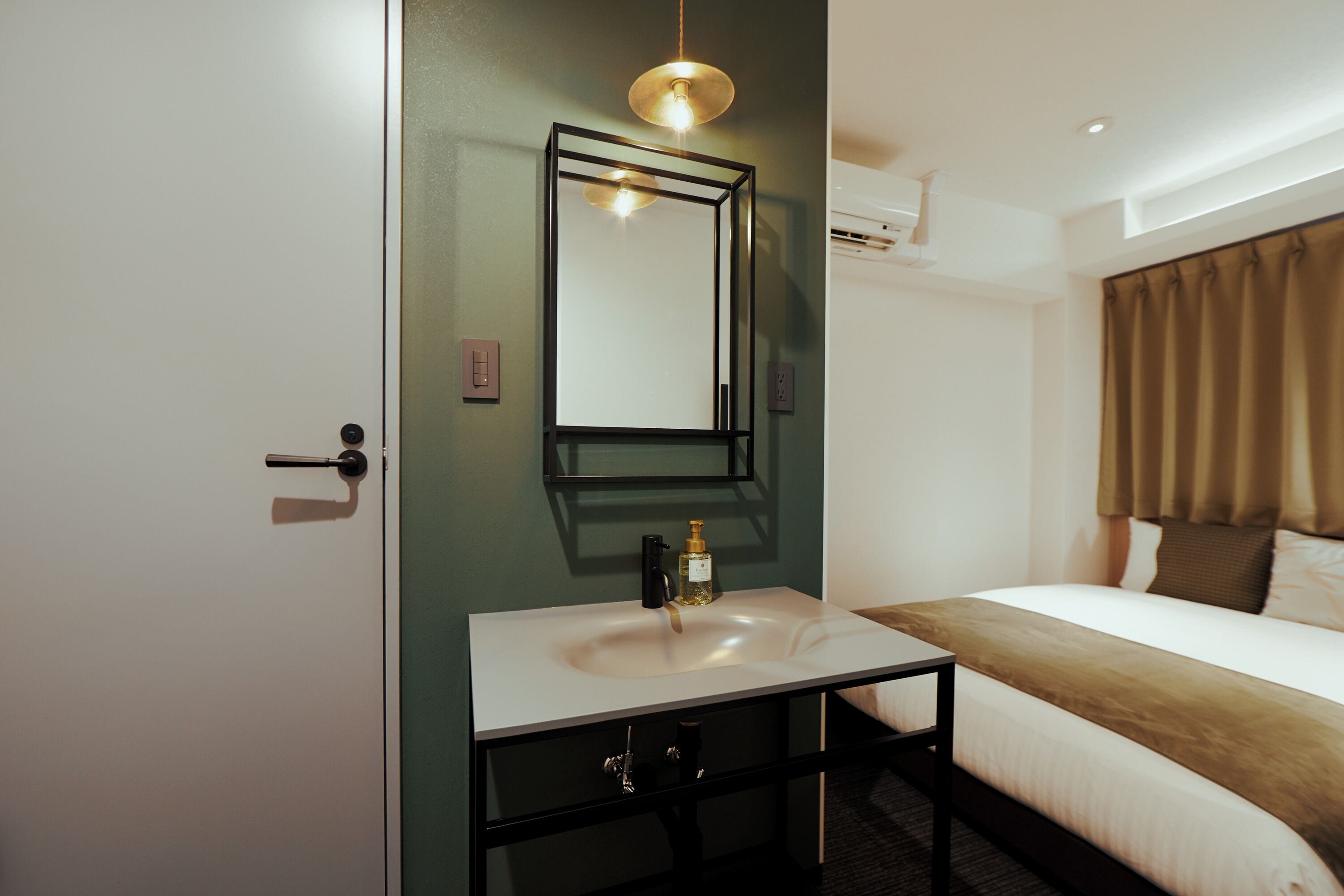 A hotel room with a white bed, a black nightstand, a white sink, a mirror, and a brown curtain.